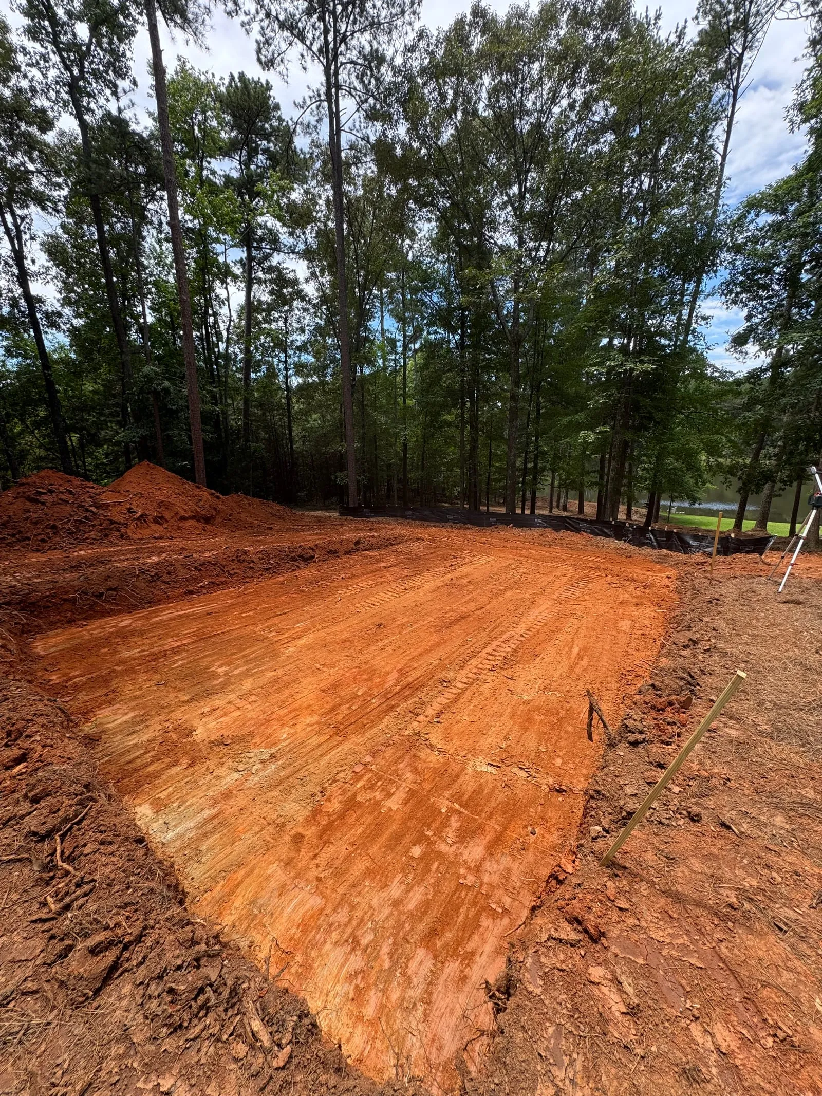 Excavated red-clay building pad on a wooded lot, with silt fence and survey stakes.